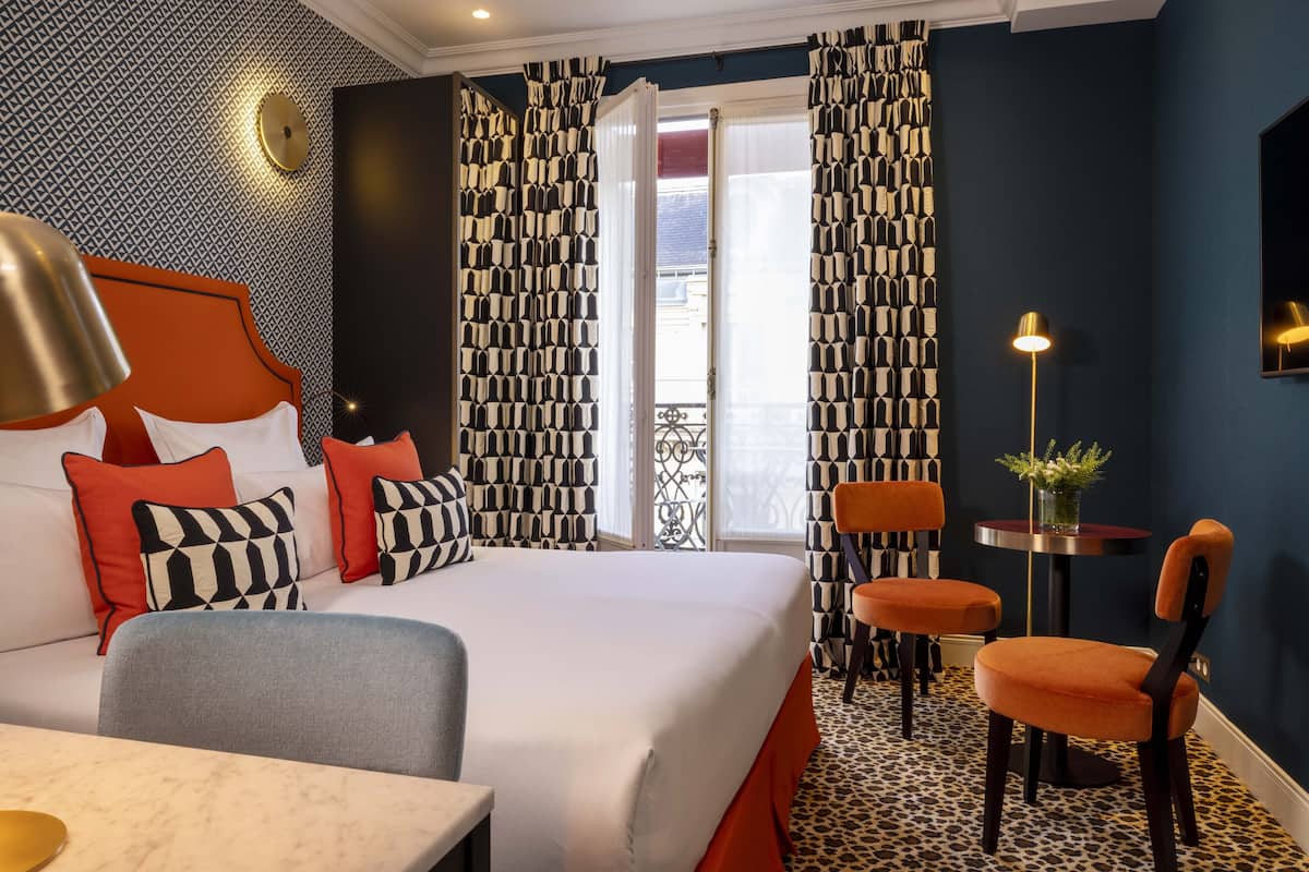 A hotel room with a large bed, two chairs, a small table, and a balcony with patterned curtains.
