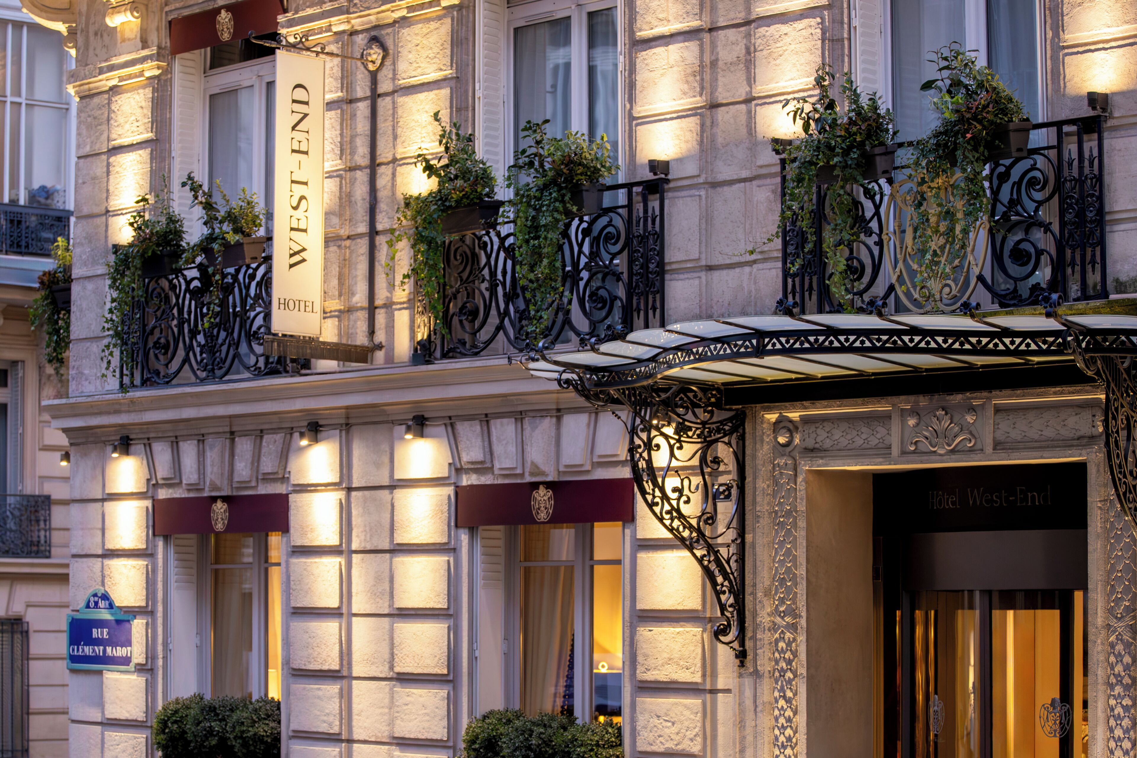 Fachada de hotel con un letrero que dice "West-End Hotel", balcones con plantas y un toldo de hierro forjado.