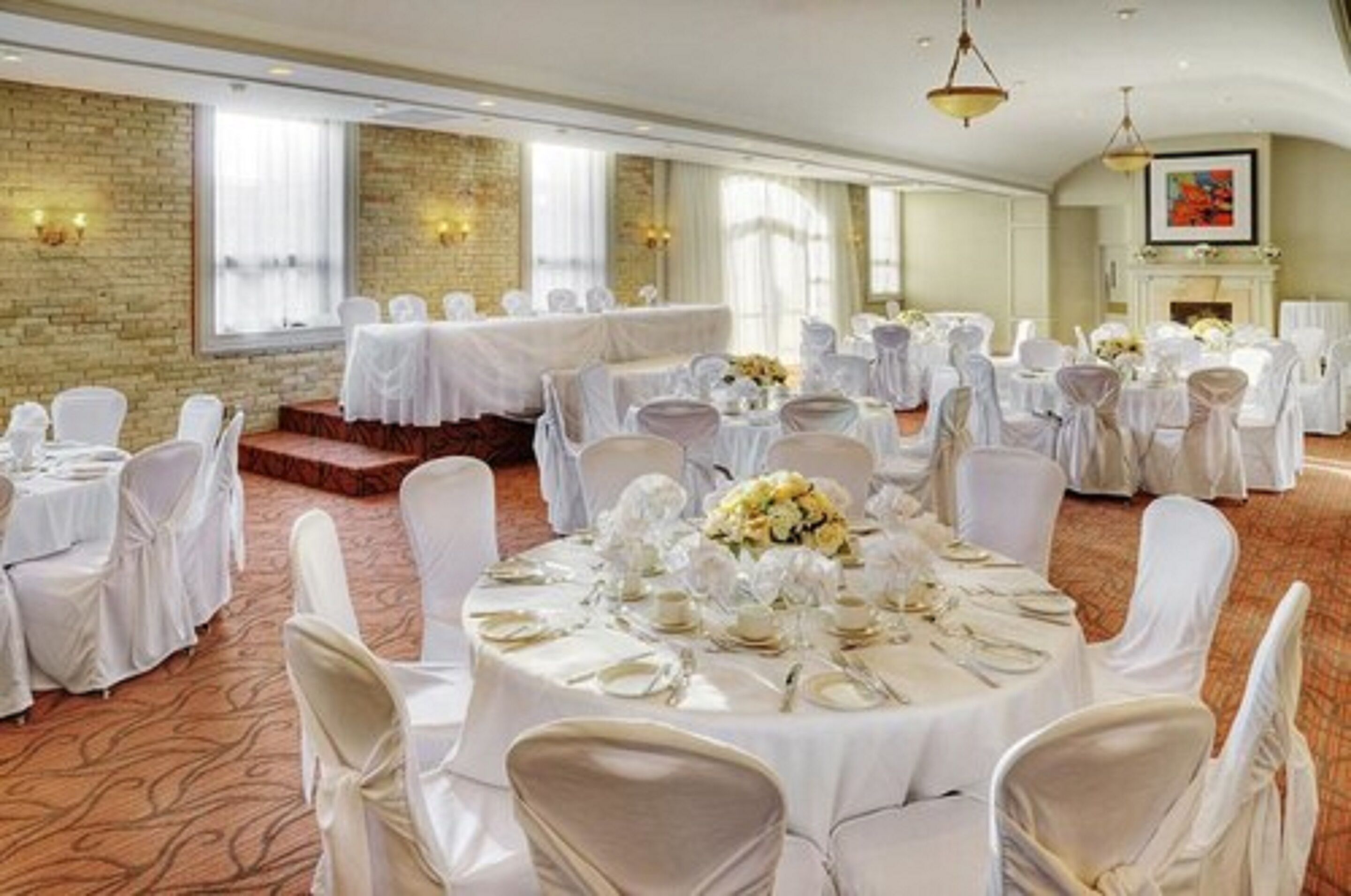 A banquet hall with round tables set for events, featuring white tablecloths and chairs, a brick wall, and large windows.
