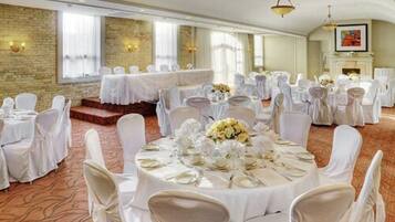 A banquet hall with round tables set for events, featuring white tablecloths and chairs, a brick wall, and large windows.