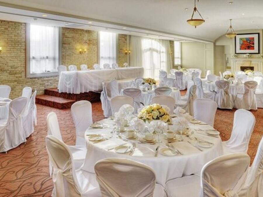 Un salón de banquetes con mesas redondas preparadas para eventos, con manteles y sillas blancos, una pared de ladrillo y ventanas amplias.