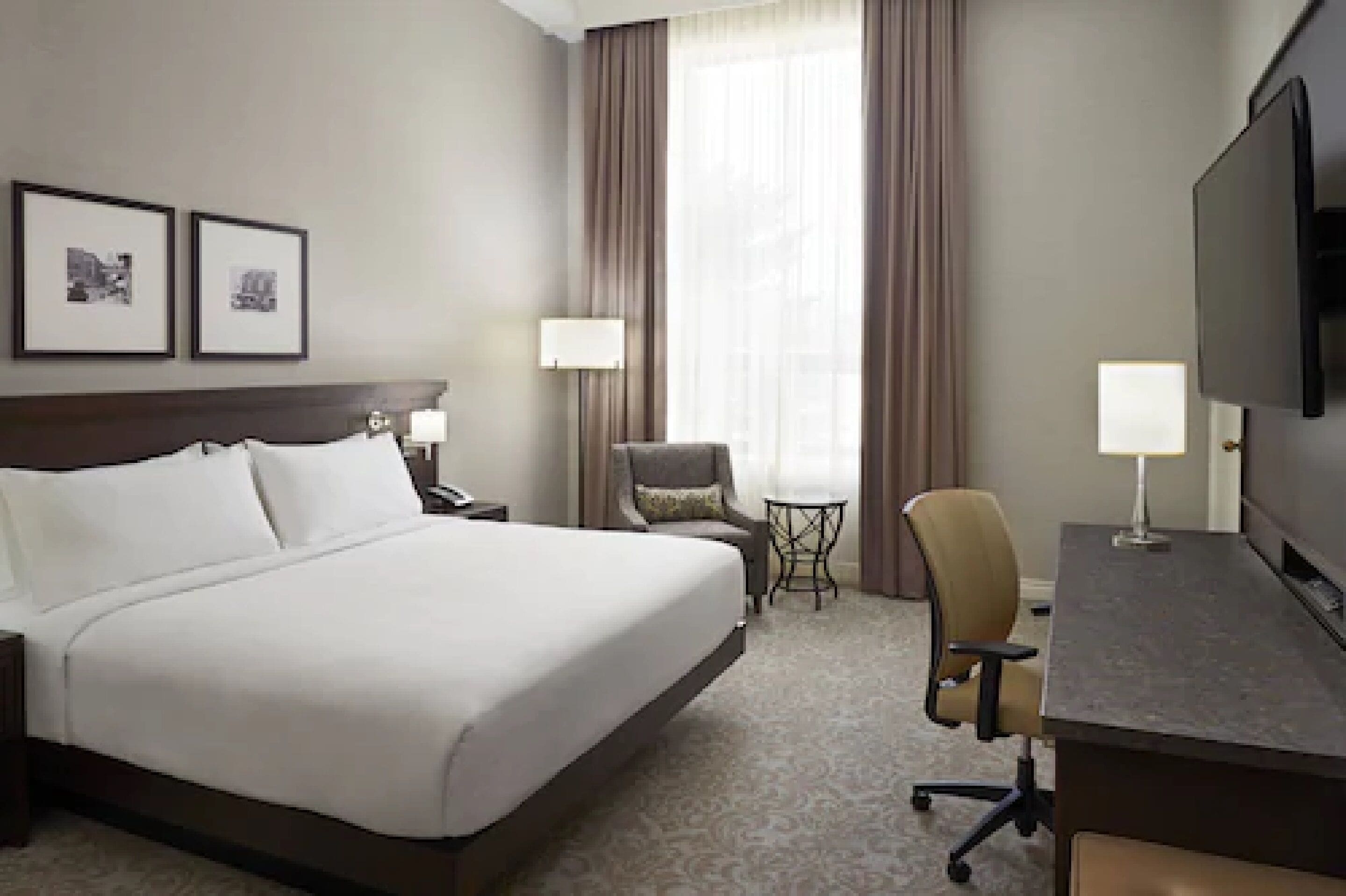 Une chambre d’hôtel avec un grand lit, un bureau avec une chaise, une télévision, deux lampes, une petite table et deux tableaux encadrés au mur.
