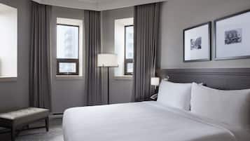 A hotel room with a large bed, two bedside lamps, a bench, and two windows with curtains.