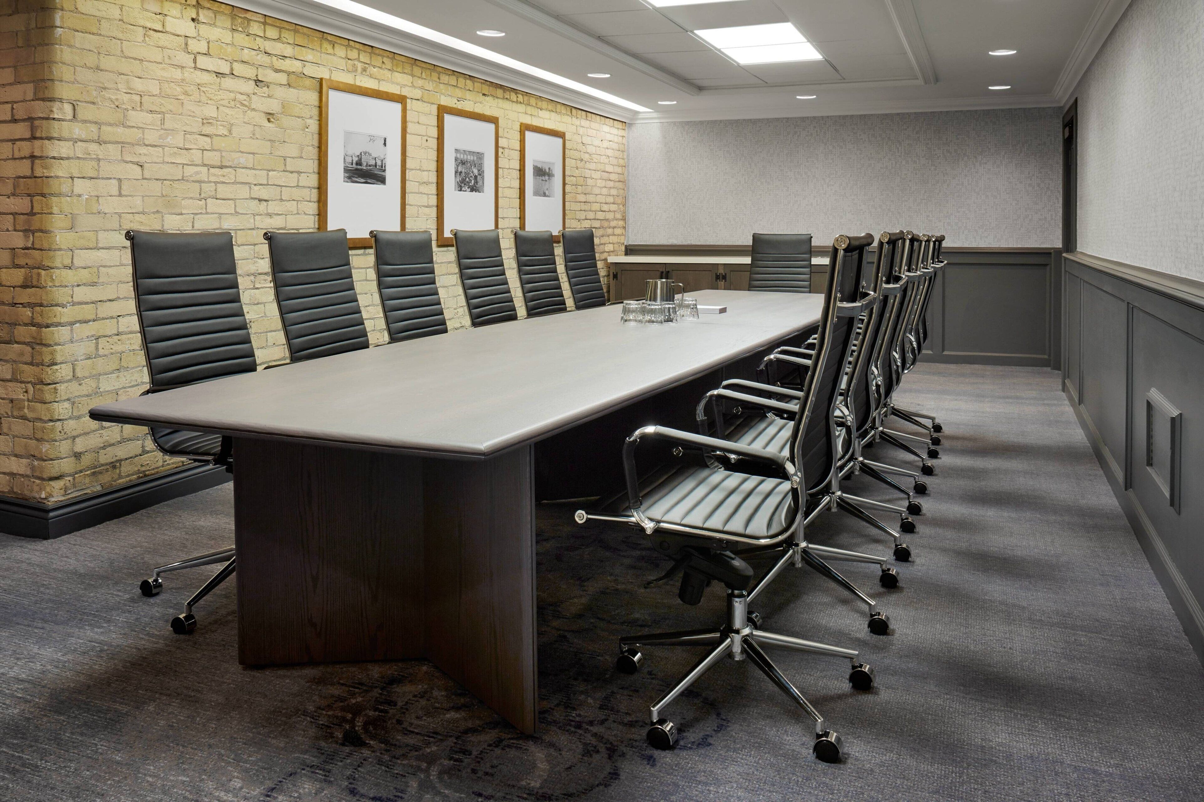 Sala de conferências com uma mesa comprida, cadeiras pretas, parede de destaque em tijolo, quadros emoldurados e piso coberto por carpete.