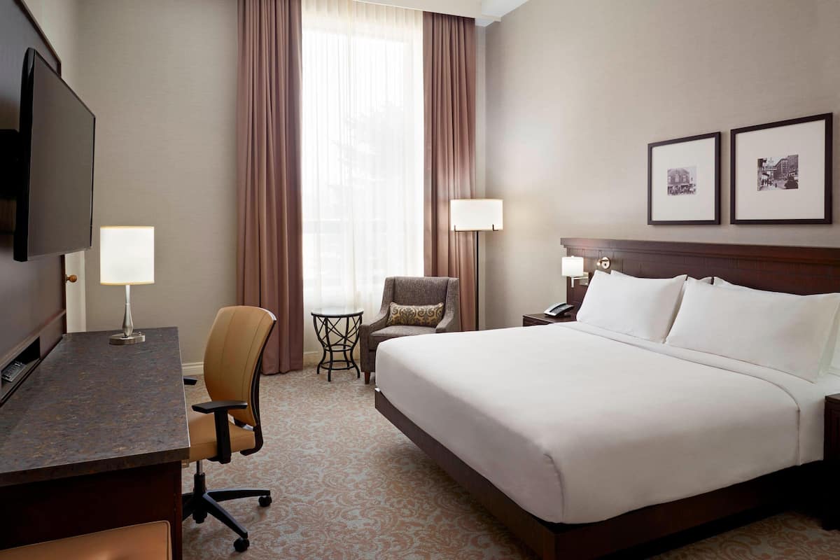 A hotel room with a bed, a desk, a chair, a TV, a lamp, and two framed pictures on the wall.