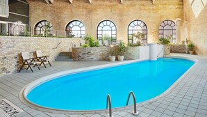 An indoor swimming pool with a brick wall, arched windows, and lounge chairs.