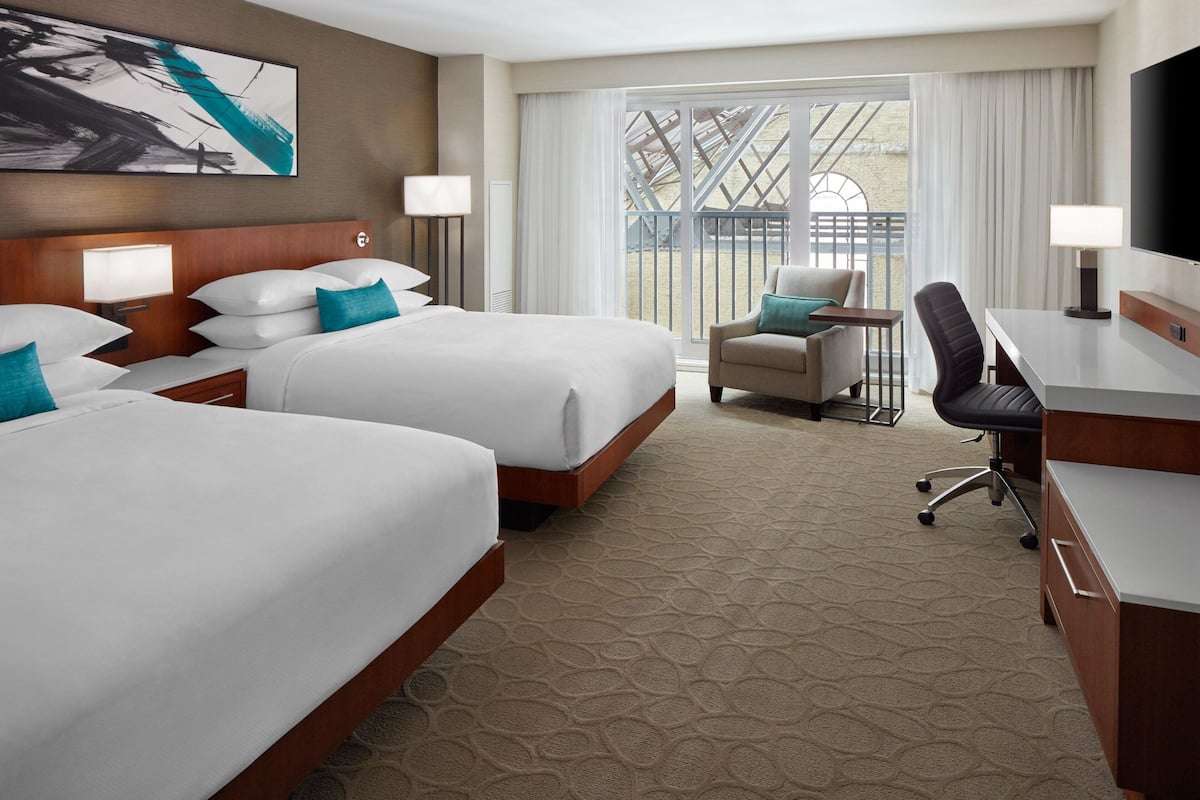 A hotel room with two beds, a desk, a chair, and a large window with a view of a building.
