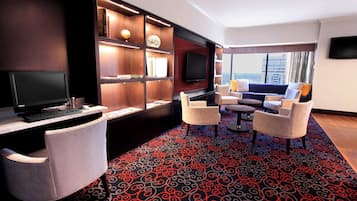 A modern hotel room with a flat-screen TV, a desk with a computer, a comfortable seating area with chairs, a patterned carpet, and a bookshelf with decorative items.