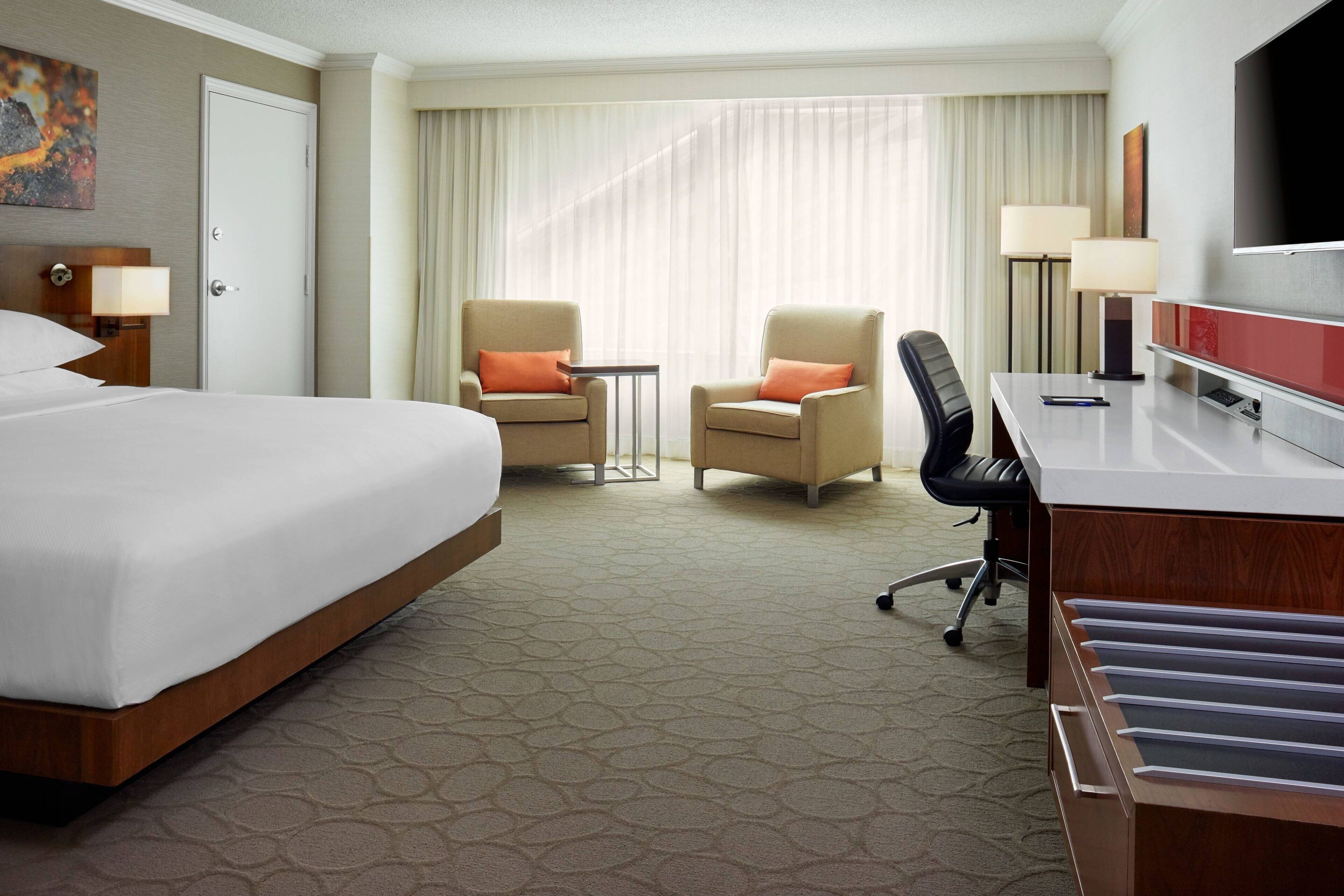 Habitación de hotel con una cama grande, dos sillones, un escritorio con silla, televisor y ventana con cortinas.