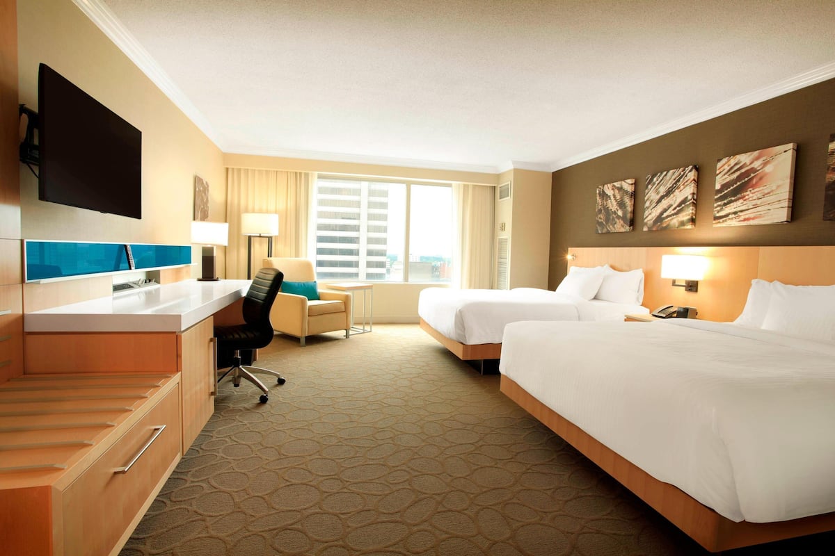 A hotel room with two beds, a desk, a chair, a TV, and artwork on the walls.