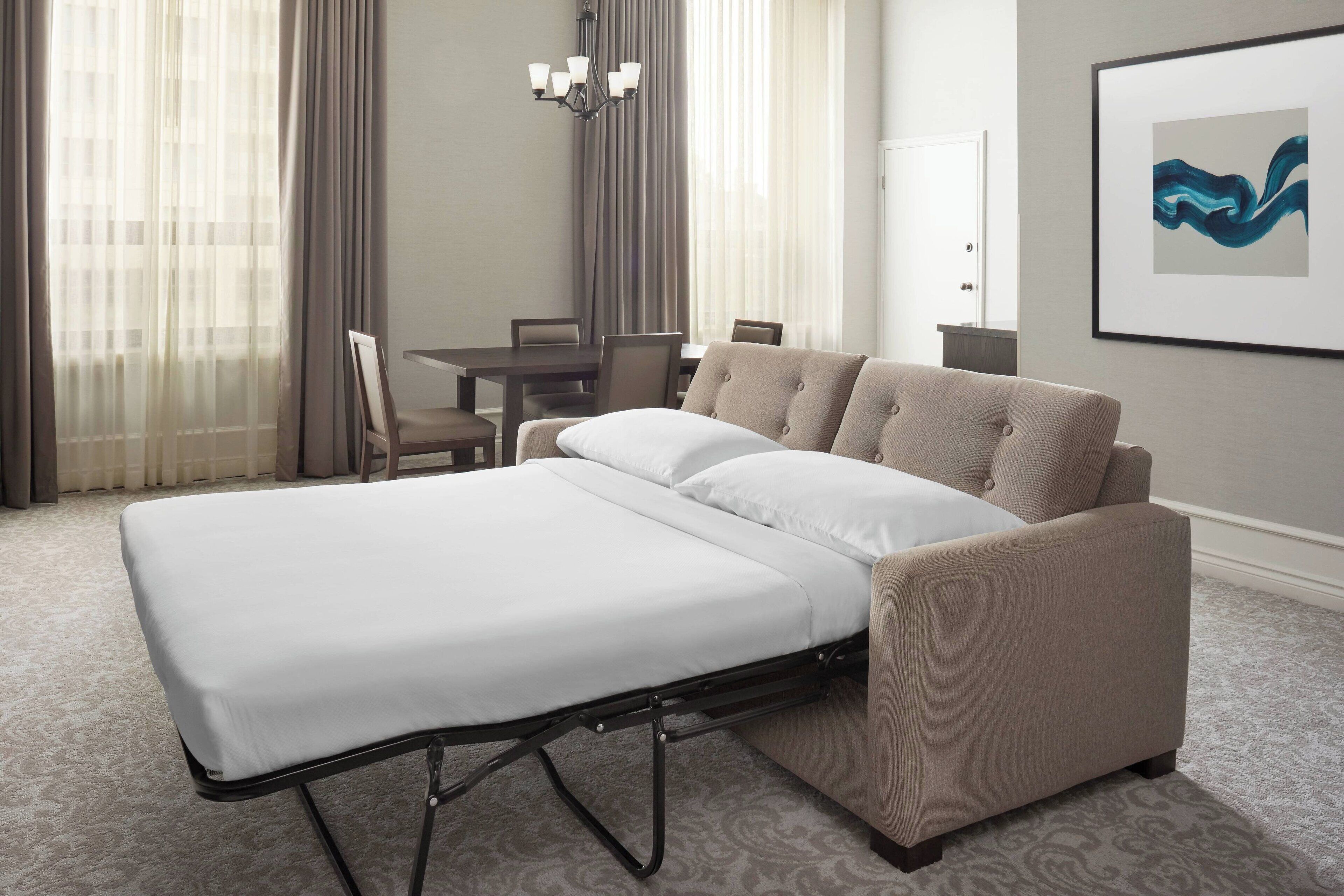 Una habitación de hotel con cama, una mesa de comedor, sillas, una araña y una obra de arte enmarcada en la pared.
