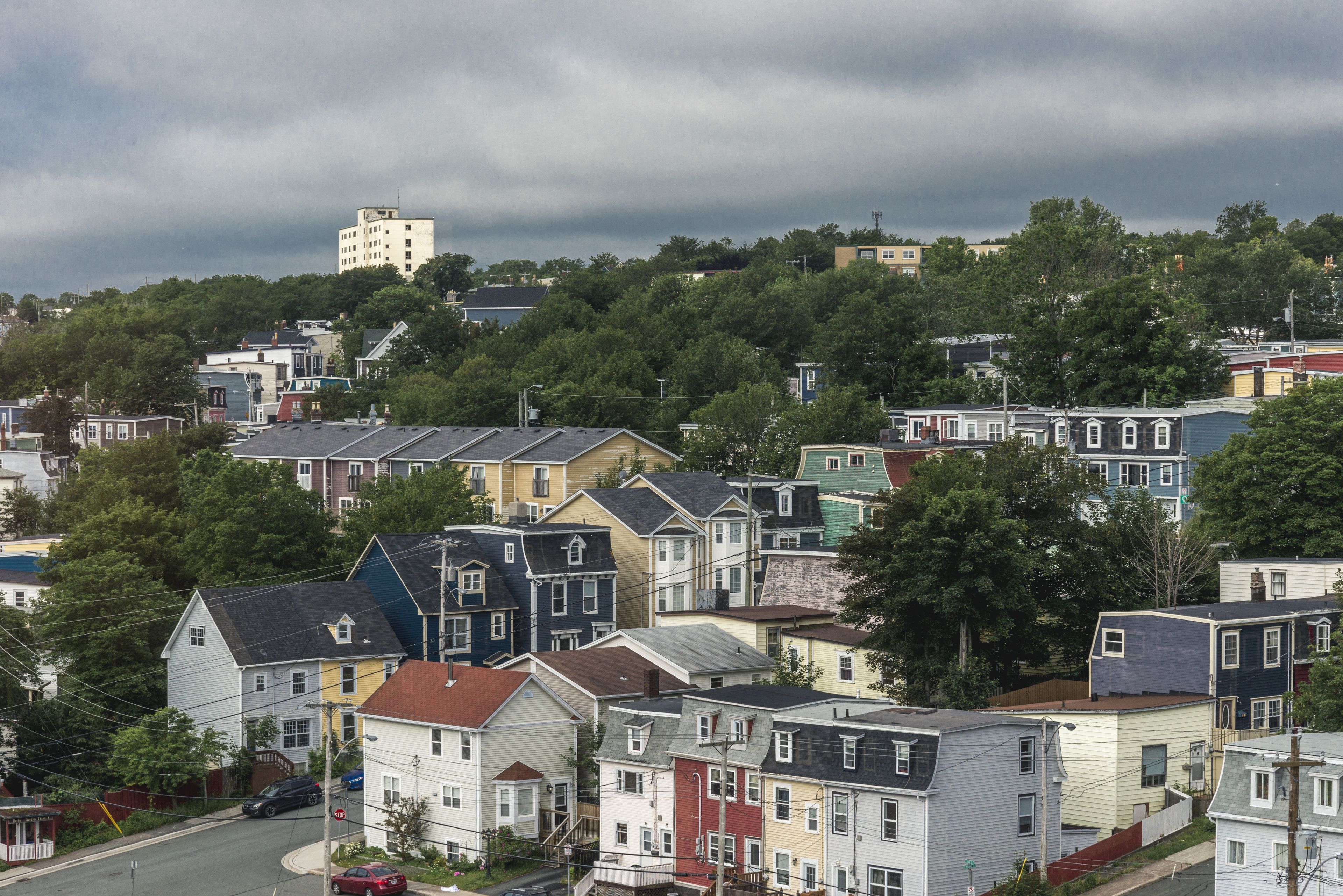A residential area with houses of various colors and architectural styles, surrounded by trees and a hill in the background.