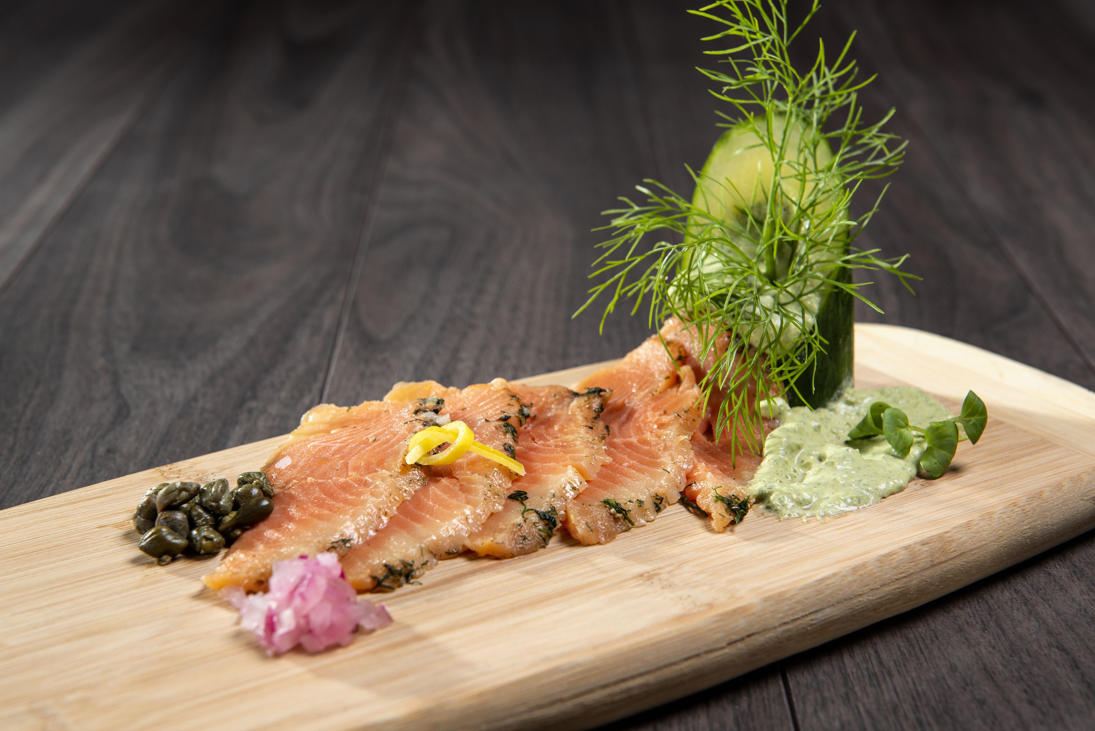 A wooden board with sliced salmon, capers, dill, lemon, and a green sauce.