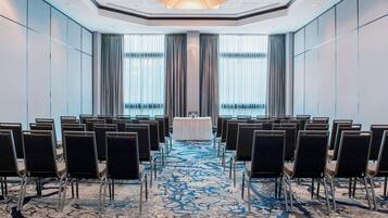 Un salón de conferencias con filas de sillas orientadas hacia una mesa al frente, ventanas amplias con cortinas y una alfombra estampada.