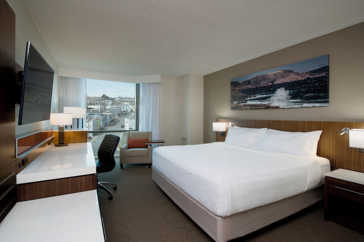 Habitación de hotel con una cama grande, un escritorio, una silla, un televisor y vista a la ciudad.