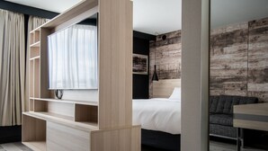 A modern hotel room with a large TV on a wooden stand, a bed with white linens, a grey sofa, and a wooden accent wall. - Delta Hotels by Marriott Sherbrooke Conference Centre (Sherbrooke)