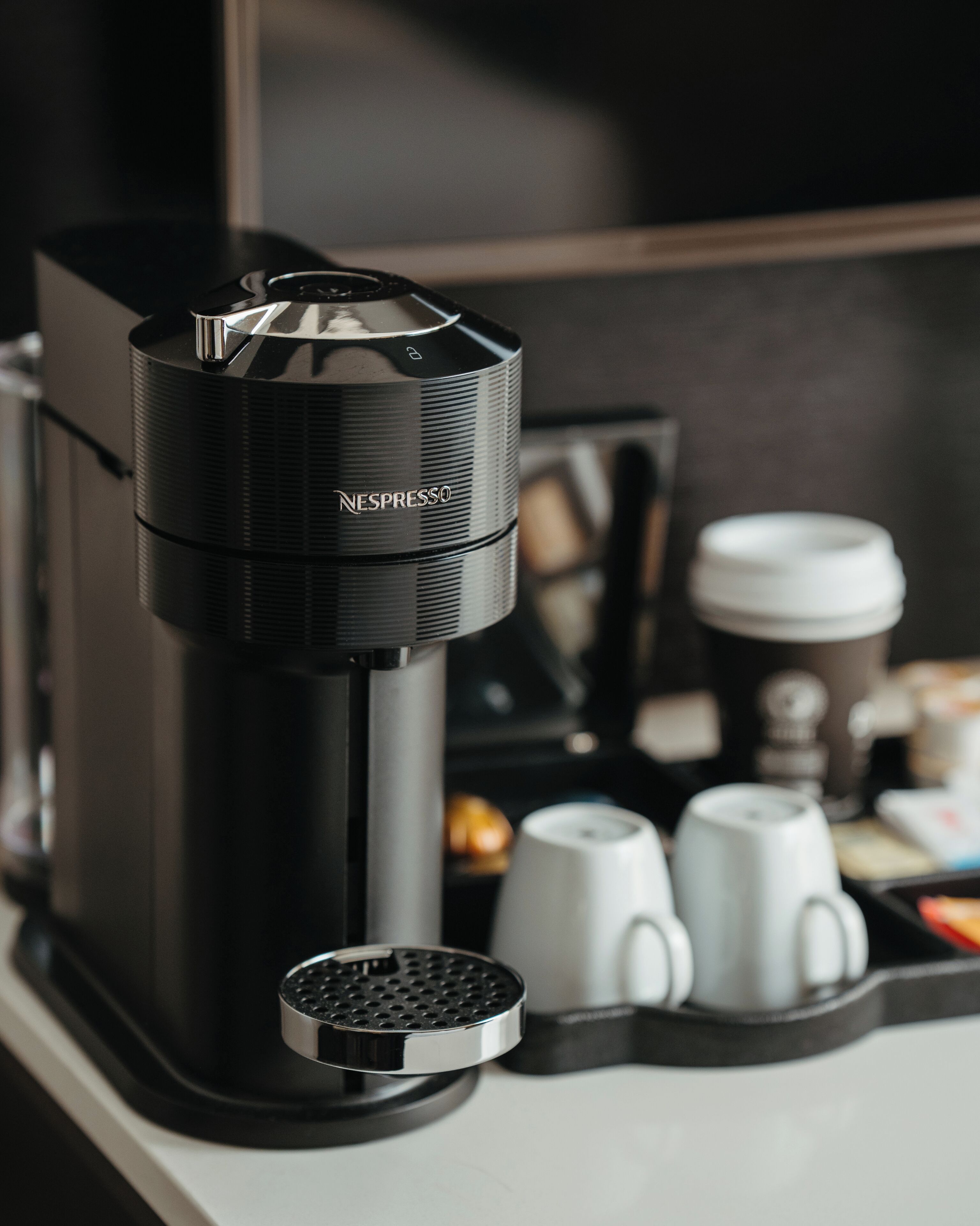Una máquina de café Nespresso con un recipiente para líquidos, dos tazas de café blancas y una jarra de café sobre una encimera.