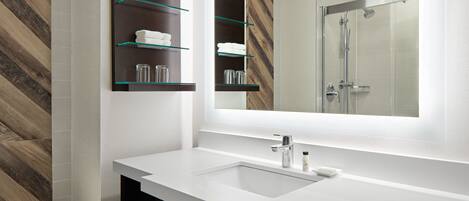 A modern bathroom with a white sink, glass shelves, and a large mirror reflecting a shower area.