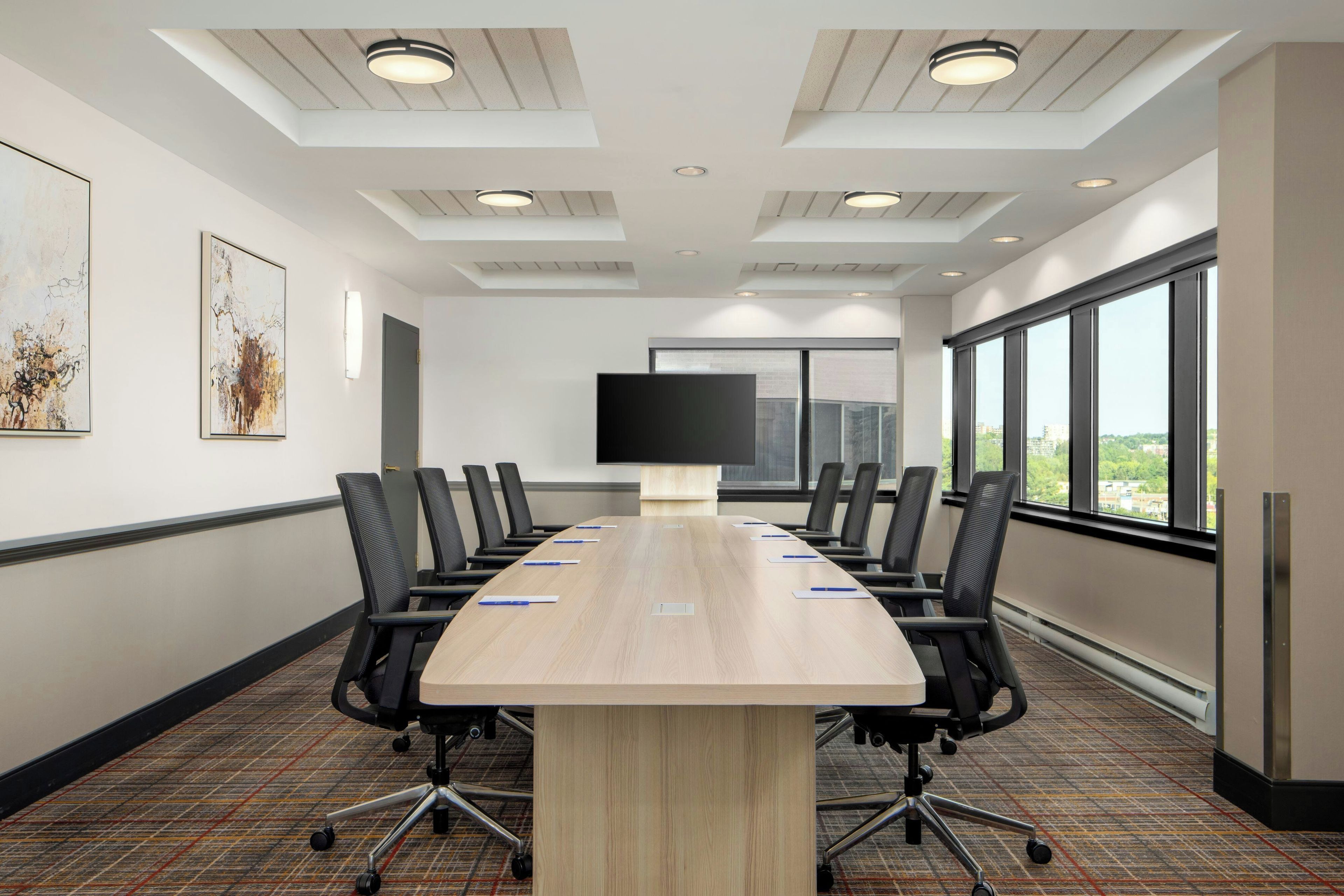 A conference room with a long table, black chairs, a flat-screen TV, and large windows.