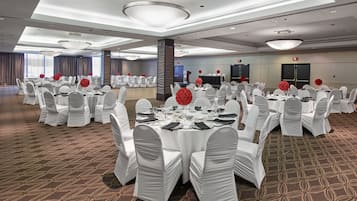 A large, well-lit banquet hall with round tables set for events, white chairs, and red floral centerpieces.