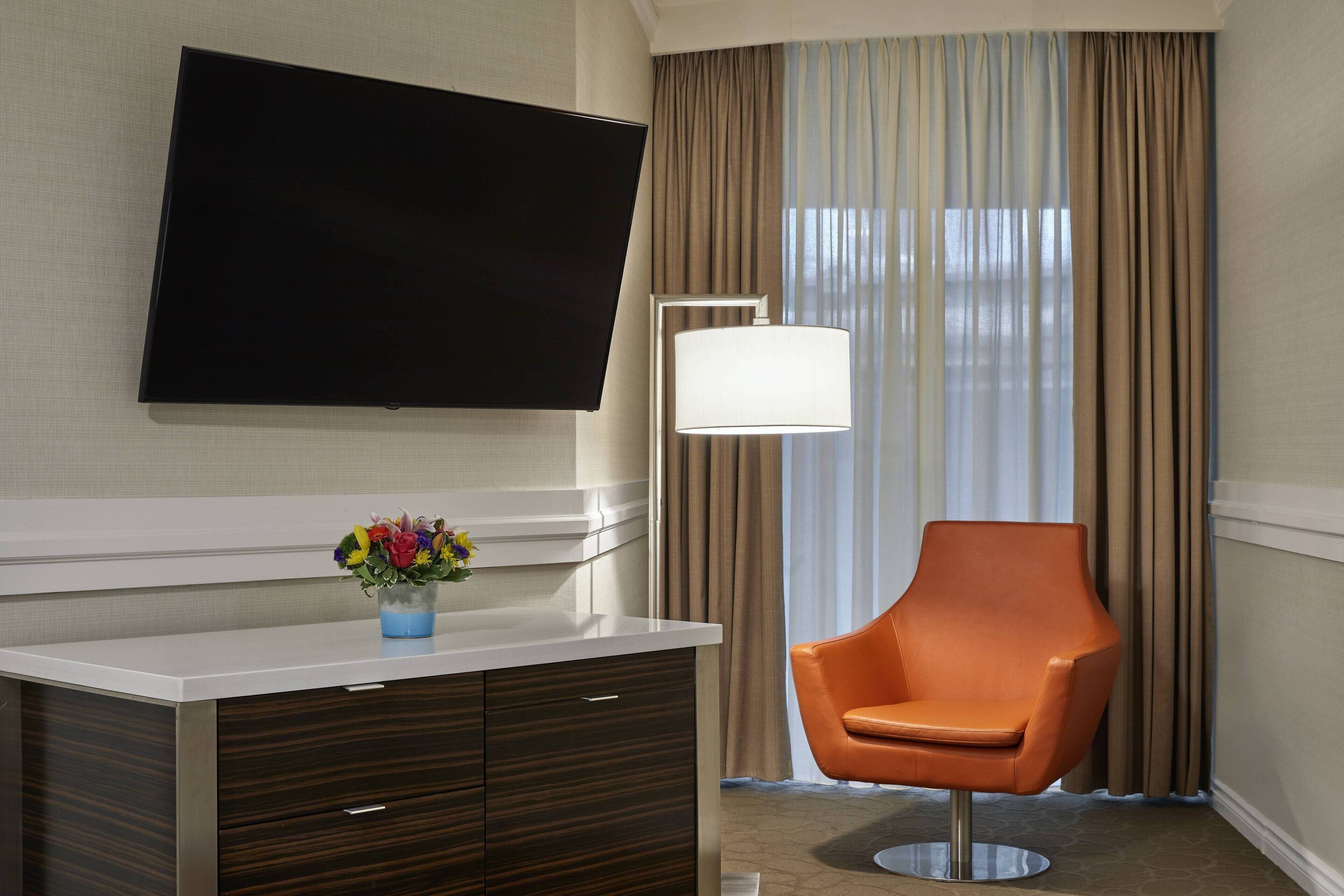 Une chambre d’hôtel dotée d’un téléviseur à écran plat fixé au mur, d’un fauteuil moderne, d’une table de chevet ornée d’un vase de fleurs et d’une grande fenêtre avec des rideaux.