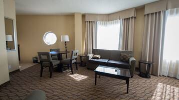 A hotel room with a sofa, two chairs, a coffee table, a round table with a lamp, and a large window with curtains.