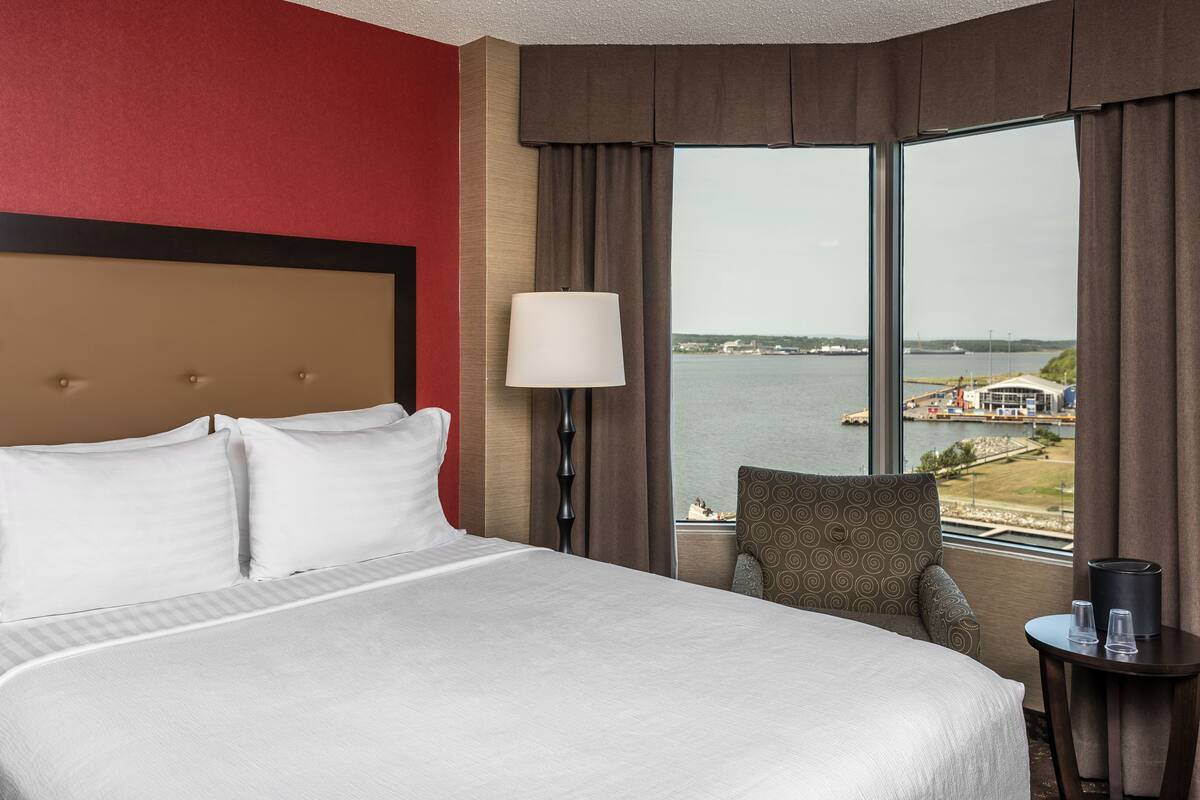 A hotel room with a large bed, a chair, a small table, a lamp, and a view of a body of water through large windows.
