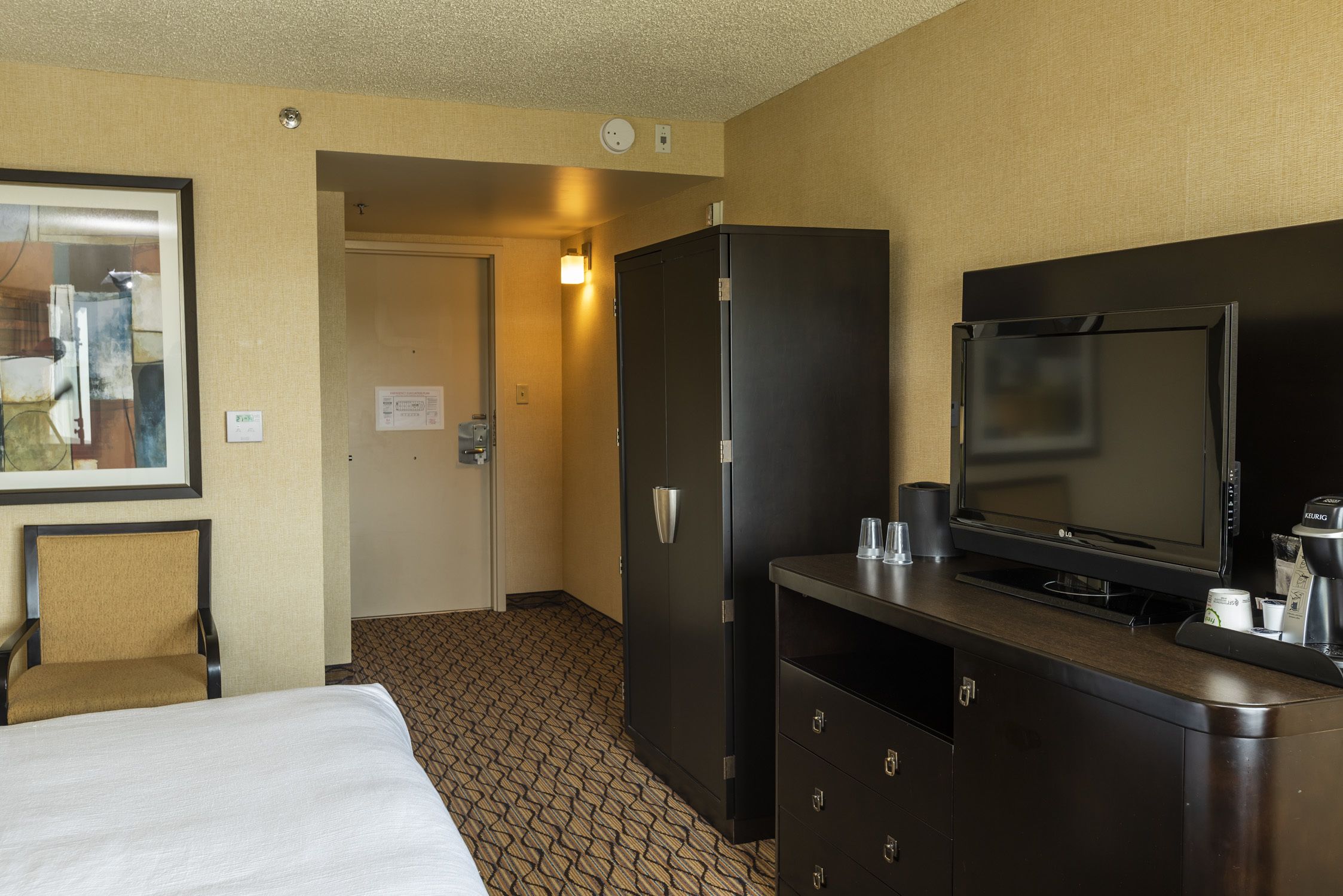 Habitación de hotel con cama, televisor, minibar y puerta que da a un pasillo.
