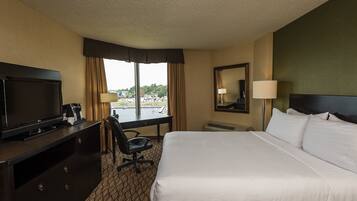 A hotel room with a large bed, a desk with a TV, a chair, a window with curtains, and a mirror.