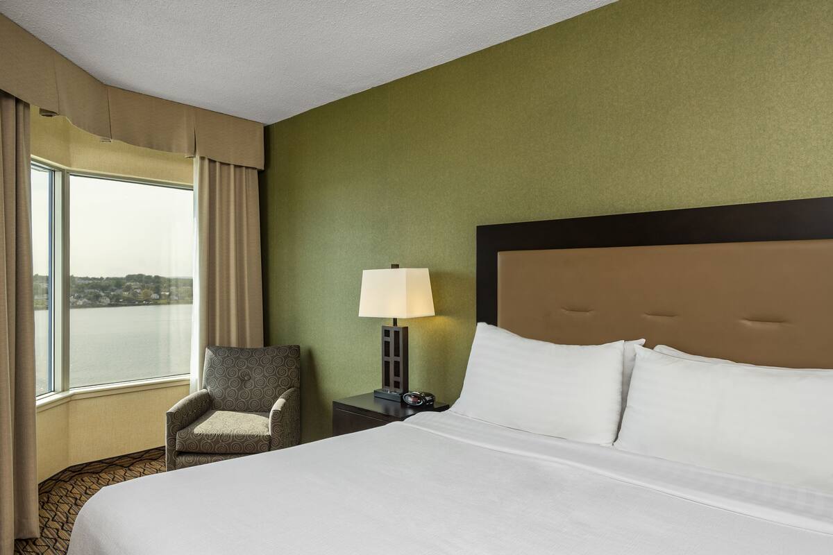 A hotel room with a large bed, a chair, a nightstand with a lamp, and a view of the water outside.
