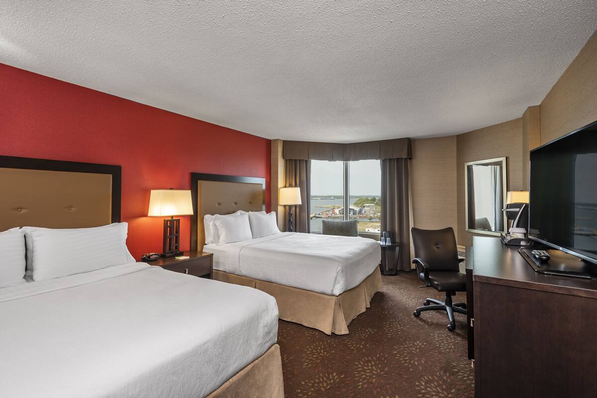 A hotel room with two beds, a desk, a chair, a TV, and a view of the city.