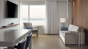 A modern hotel room with a flat-screen TV, a dining area with a table and chairs, a sofa, and a large window offering a view of the cityscape. - Delta Hotels by Marriott Trois Rivieres Conference Centre (Trois-Rivieres)