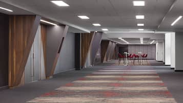 A long, narrow hallway with a patterned carpet, wooden paneling on the walls, and a row of red chairs at the end.