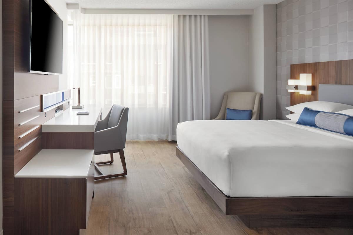 Une chambre d’hôtel moderne dotée d’une télévision à écran plat, d’un bureau avec une chaise, d’un lit avec des oreillers et d’une grande fenêtre avec des rideaux.