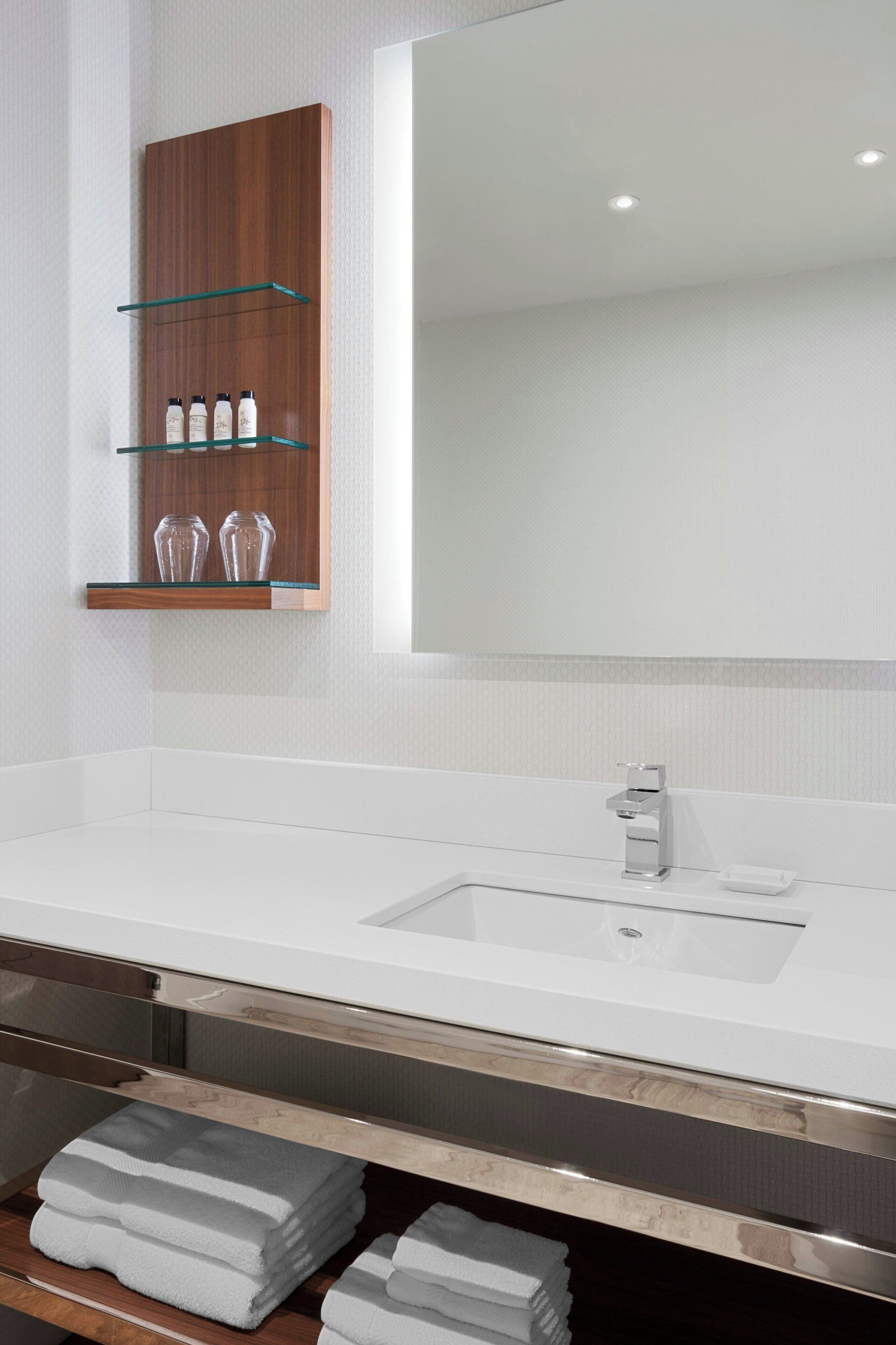 Un baño moderno con un lavamanos blanco, un espejo grande y un mueble de tocador de madera con estantes de vidrio.