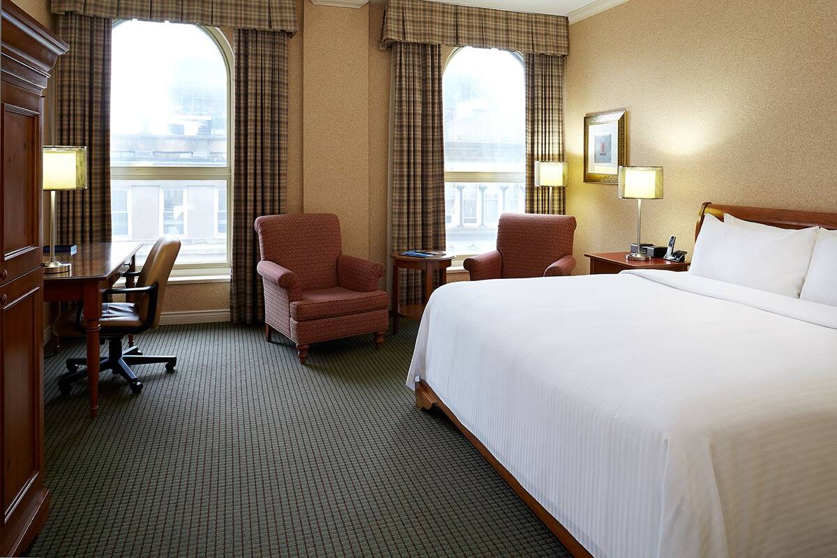 A hotel room with a large bed, two armchairs, a desk, and a view of the city through large windows.