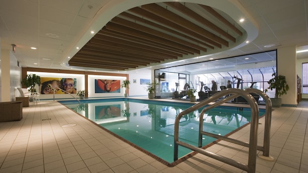 Una piscina interior con vista despejada del techo, suelo de baldosas y zona de estar junto a la piscina con sillas y plantas en macetas.