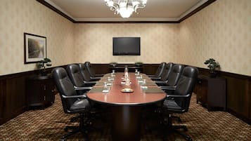 Une salle de conférence avec une longue table, des fauteuils en cuir noir, un lustre, un téléviseur à écran plat et une moquette à motifs.