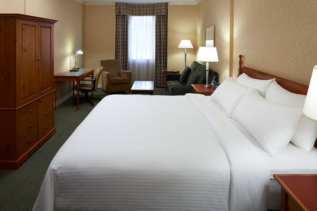 A hotel room with a large bed, a wooden wardrobe, a desk with a lamp, a chair, and a small table.