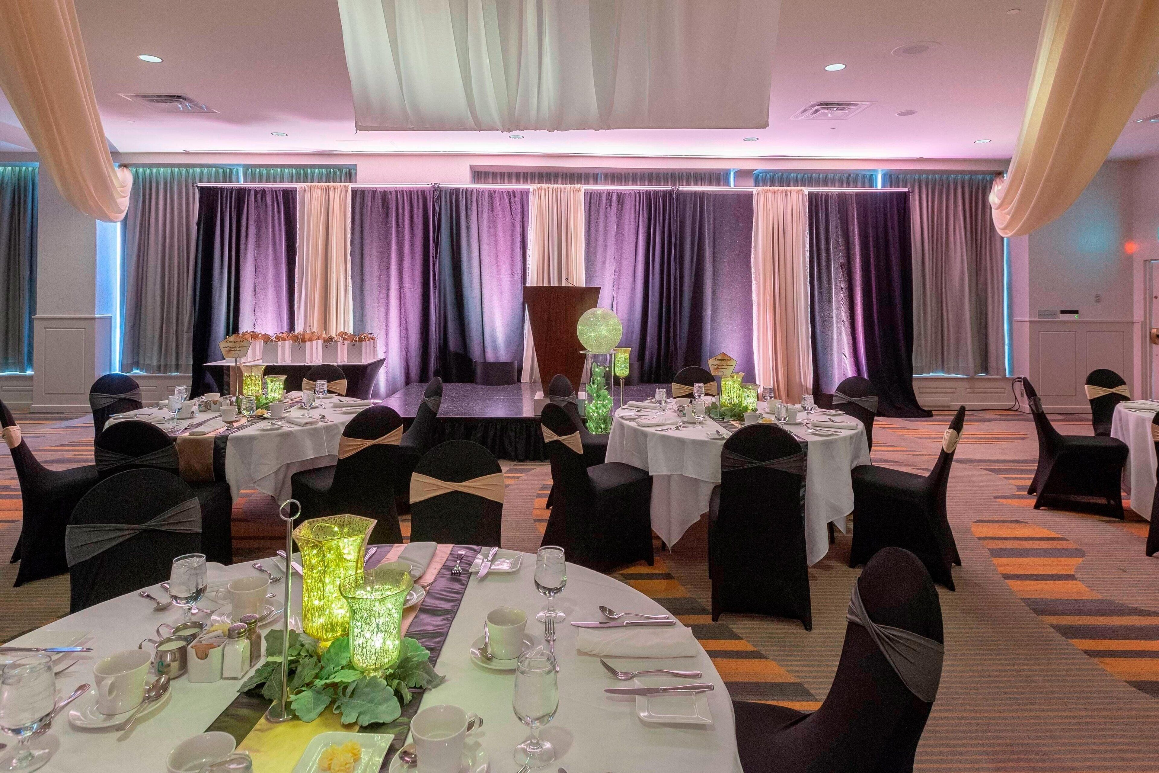A banquet hall with round tables set for dining, draped curtains, and a stage in the background.