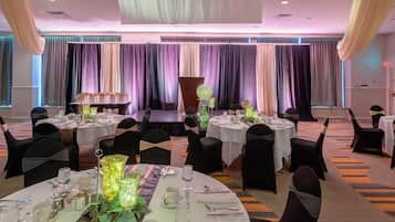 A banquet hall with round tables set for dining, draped curtains, and a stage in the background.