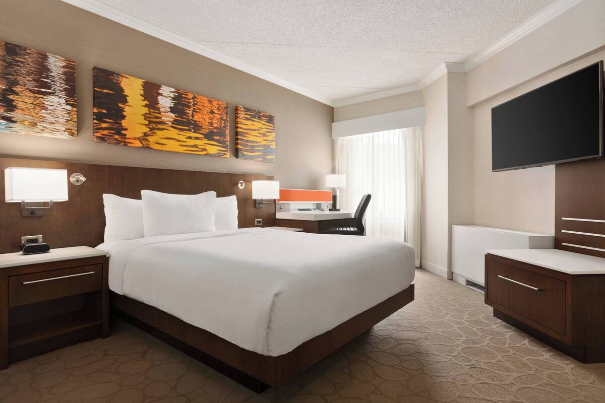 Una habitación de hotel con una cama grande, mesitas de noche, un escritorio con una silla, un televisor de pantalla plana y una obra de arte en la pared.