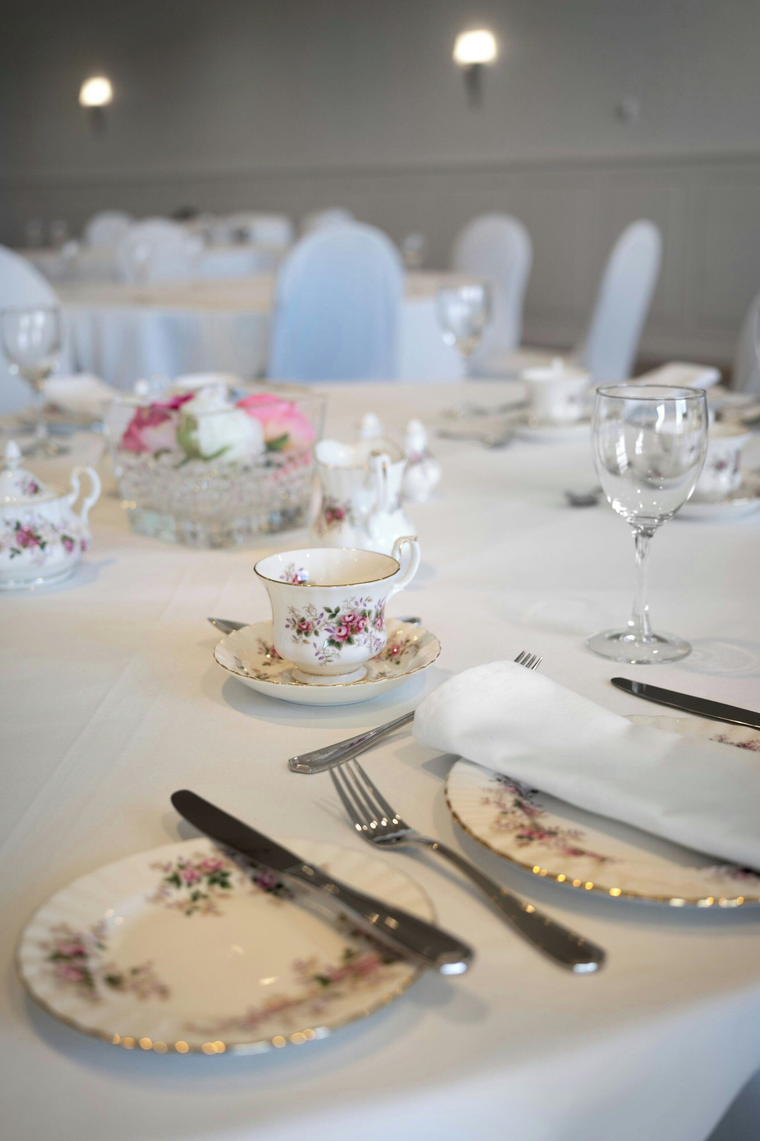 A table set with floral-patterned tableware, including a teapot, cups, and plates, with a clear glass wine glass on the table.