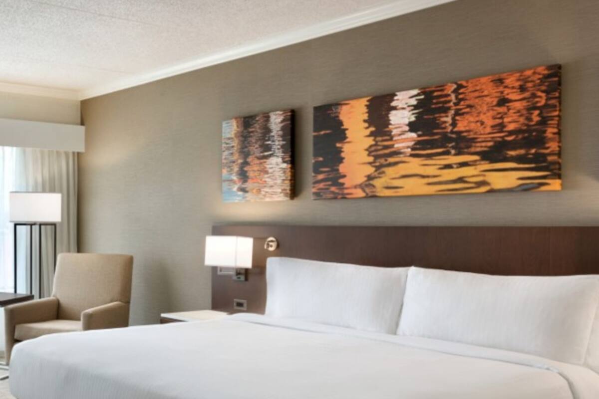 Habitación de hotel con una cama grande, una silla, una mesita de noche con lámpara y dos cuadros en la pared.
