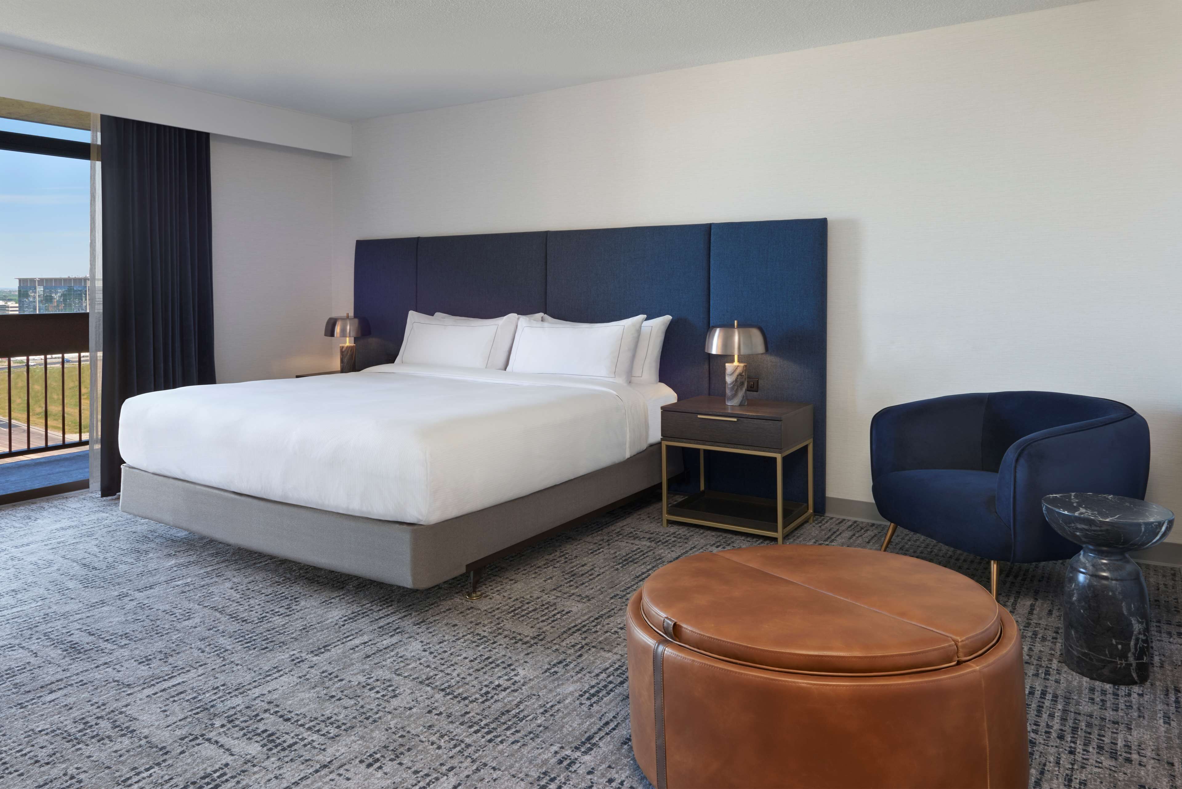 A hotel room with a large bed, a nightstand with lamps, a blue chair, and a brown ottoman.