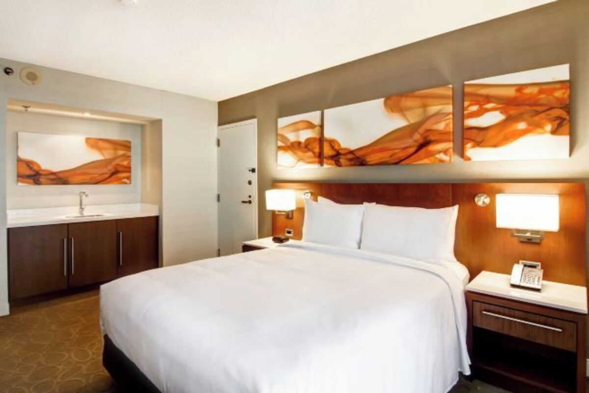 A hotel room with a large bed, a bedside table with a lamp, a nightstand with a phone, a bathroom with a sink and mirror, and abstract wall art.