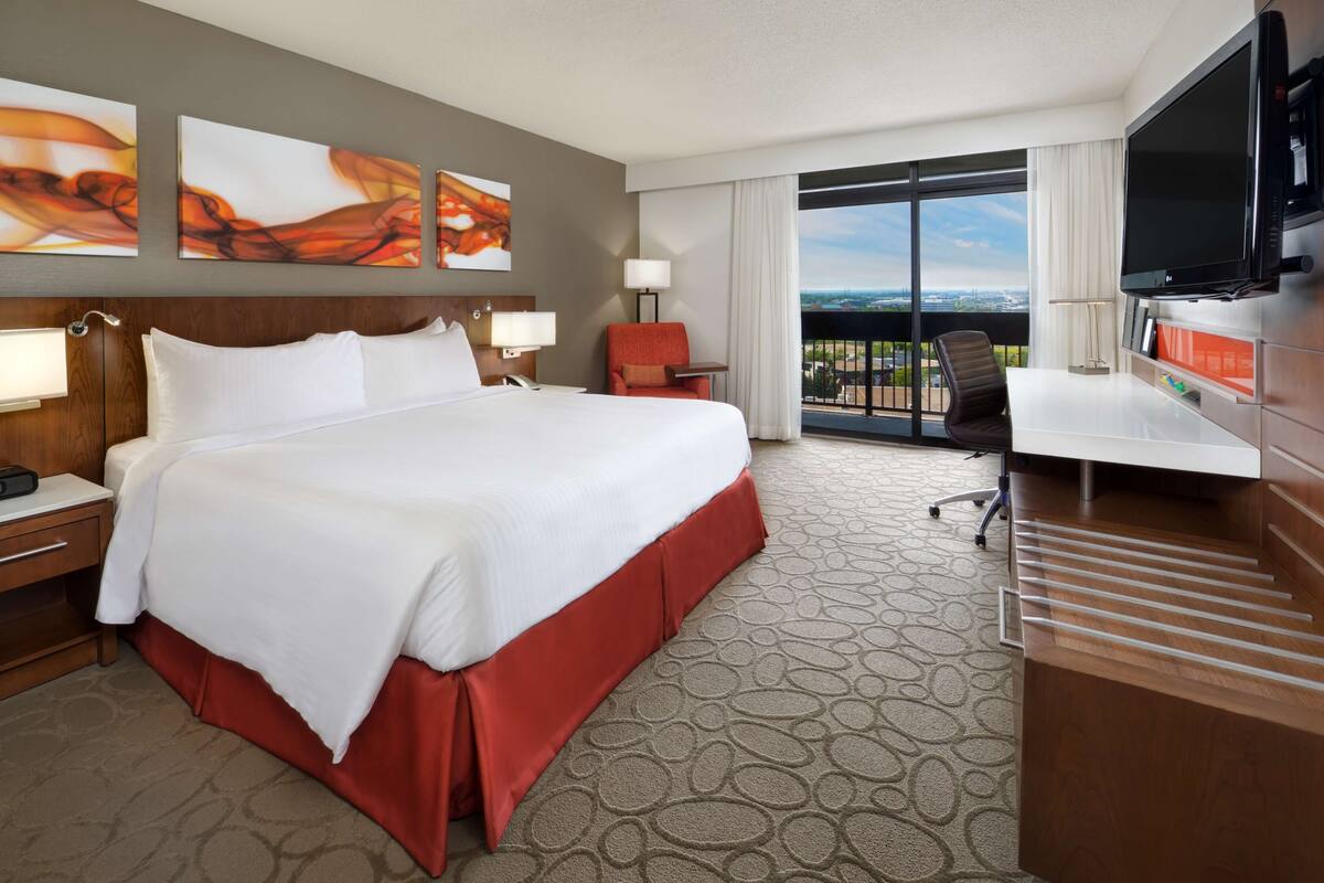A hotel room with a large bed, a desk, a TV, and a balcony with a city view.