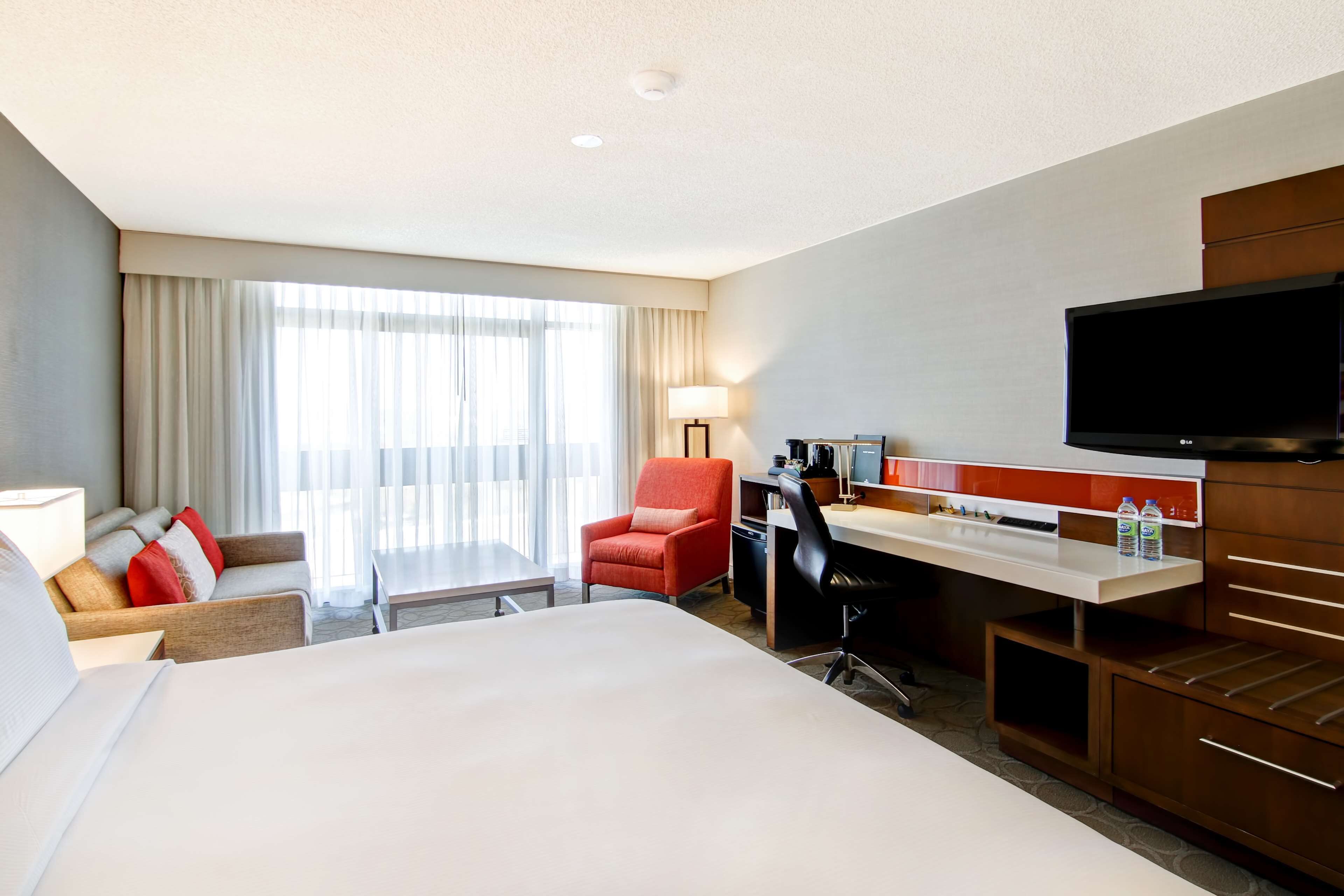 A hotel room with a large bed, a desk with a computer, a chair, a TV, and a window with curtains.