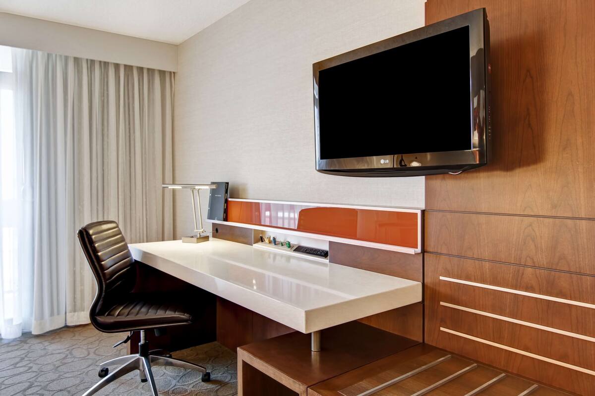 A hotel room with a flat-screen TV mounted on the wall, a white desk with a chair, and a wooden cabinet with drawers.