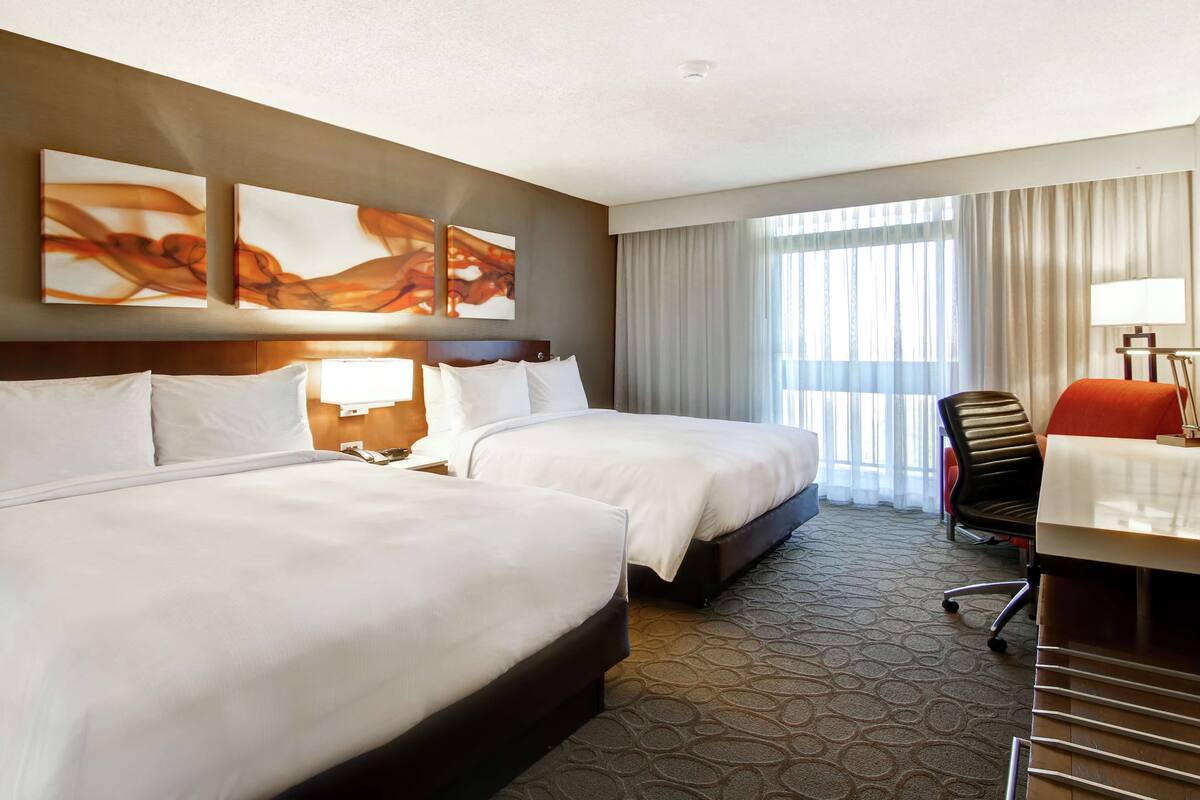 A hotel room with two beds, a desk with a chair, and a large window with curtains.